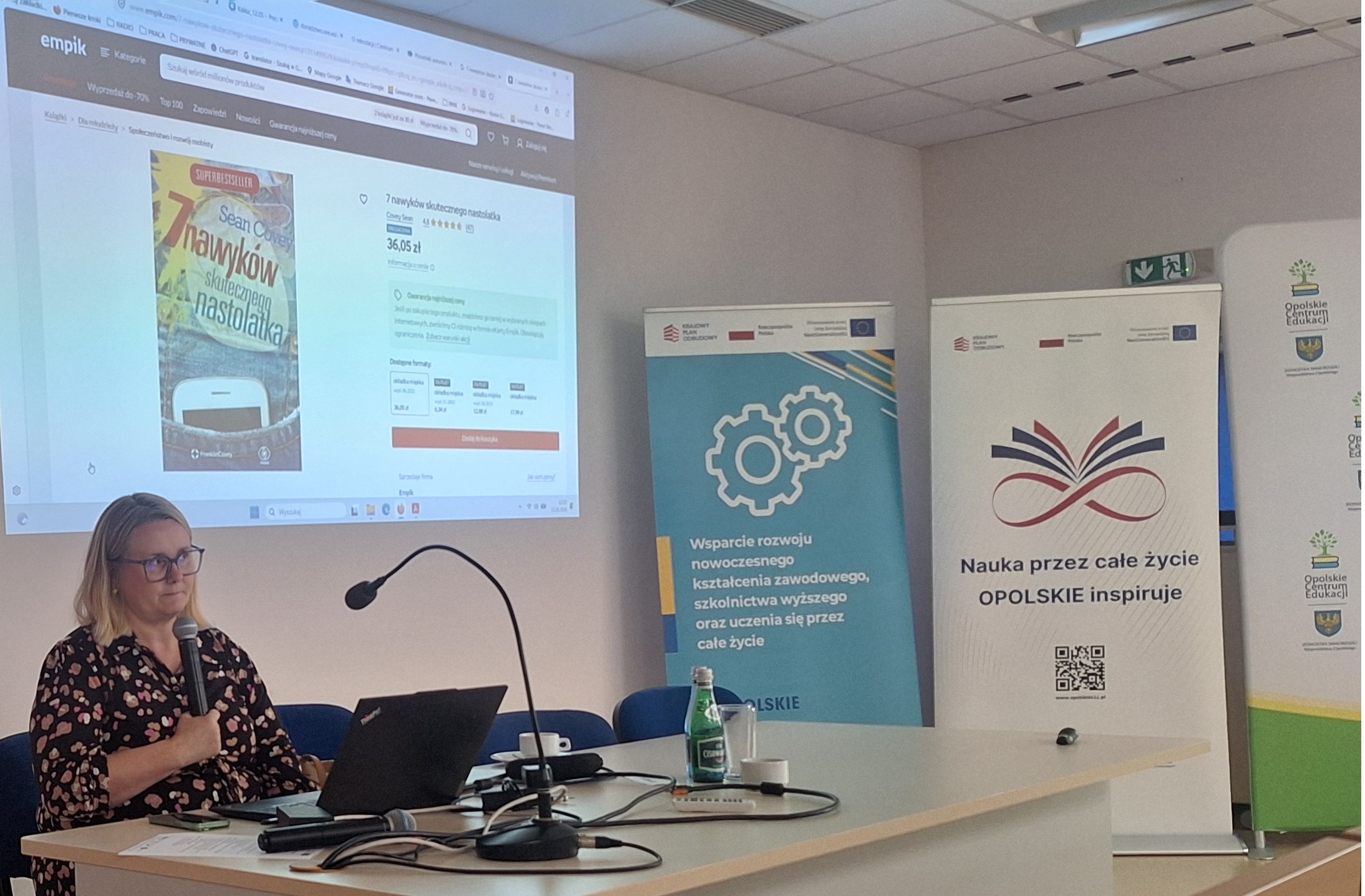 Prelegentka prezentująca slajdy podczas spotkania, widoczne banery informacyjne i ekran projekcyjny