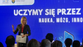 Kobieta w ciemnym garniturze w paski stoi na scenie i przemawia, trzymając w ręku podkładkę z dokumentami. W tle widoczny jest jaskrawoniebieski ekran z dużym białym napisem „UCZYMY SIĘ PRZEZ... NAUKA, MÓZG, INNOWACJE...”. W lewym górnym rogu ekranu umieszczono logotypy organizatorów, w tym Opolskiego Centrum Rozwoju Gospodarki. Na pierwszym planie widać głowy uczestników wydarzenia siedzących na widowni.