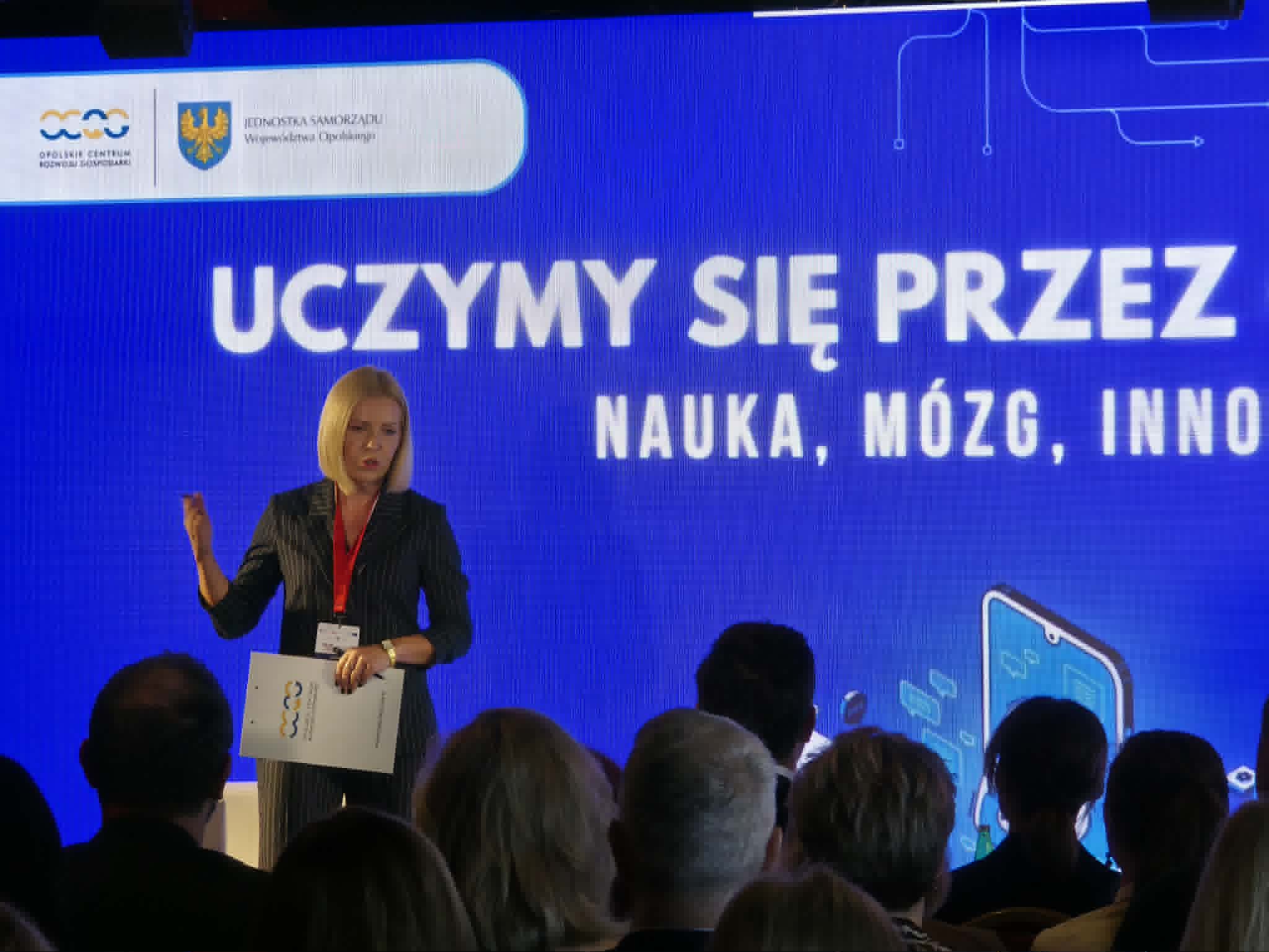 Kobieta w ciemnym garniturze w paski stoi na scenie i przemawia, trzymając w ręku podkładkę z dokumentami. W tle widoczny jest jaskrawoniebieski ekran z dużym białym napisem „UCZYMY SIĘ PRZEZ... NAUKA, MÓZG, INNOWACJE...”. W lewym górnym rogu ekranu umieszczono logotypy organizatorów, w tym Opolskiego Centrum Rozwoju Gospodarki. Na pierwszym planie widać głowy uczestników wydarzenia siedzących na widowni.