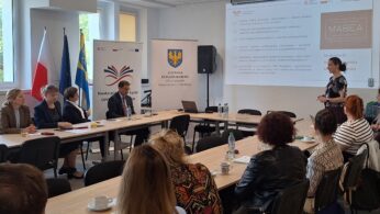 Spotkanie panelowe w sali konferencyjnej, przy stole siedzą prowadzący, a uczestnicy oglądają prezentację na ekranie.