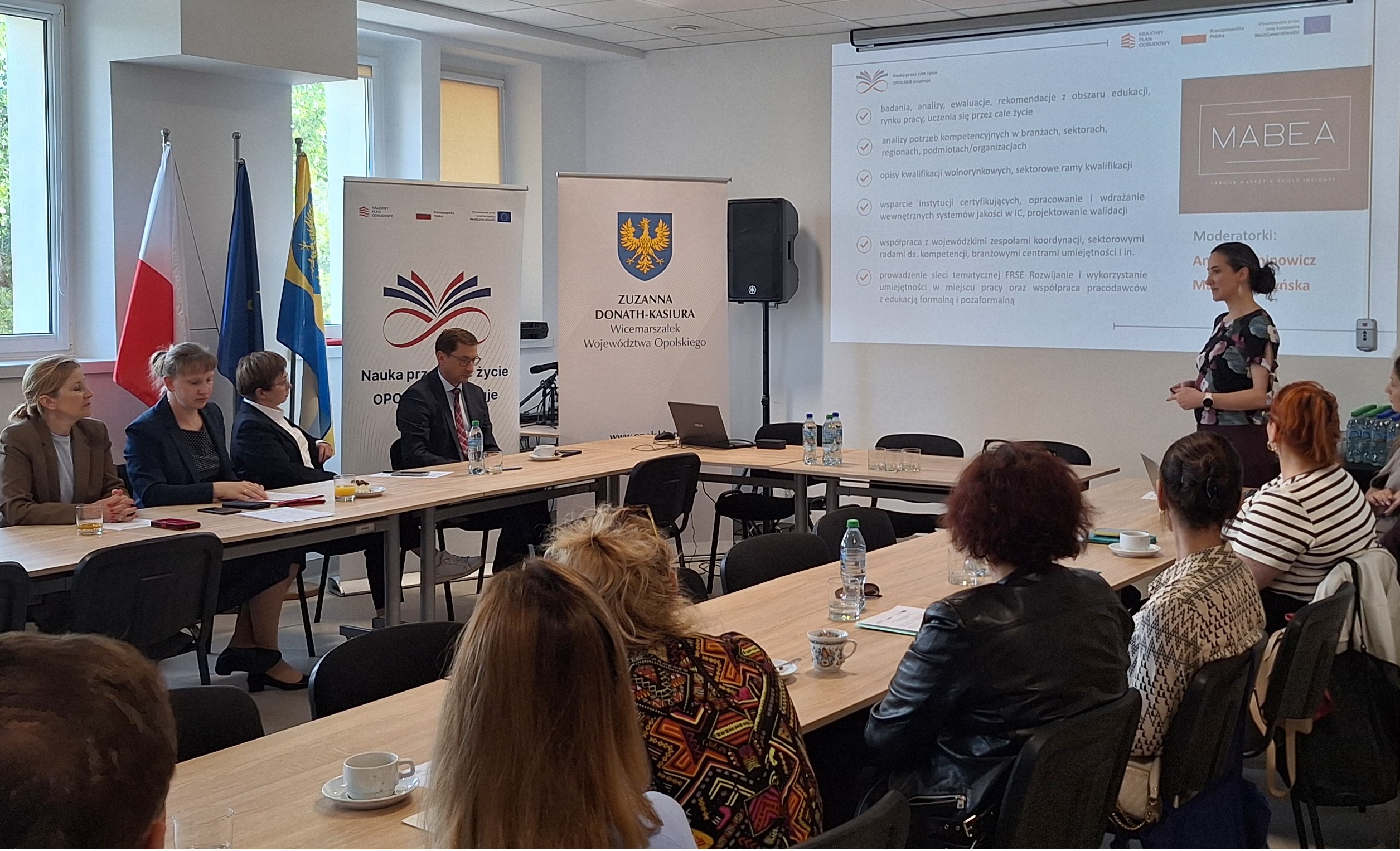 Spotkanie panelowe w sali konferencyjnej, przy stole siedzą prowadzący, a uczestnicy oglądają prezentację na ekranie.