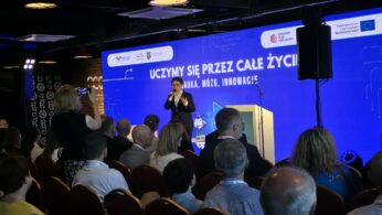 Kobieta stoi na scenie za mównicą i przemawia do mikrofonu podczas oficjalnego wydarzenia. Tło stanowi ogromny niebieski ekran z hasłem „UCZYMY SIĘ PRZEZ CAŁE ŻYCIE! NAUKA, MÓZG, INNOWACJE”. Na pierwszym planie widoczni są uczestnicy konferencji siedzący w rzędach na krzesłach z bordową tapicerką. Po lewej stronie jedna z osób na widowni rejestruje wystąpienie za pomocą telefonu komórkowego.