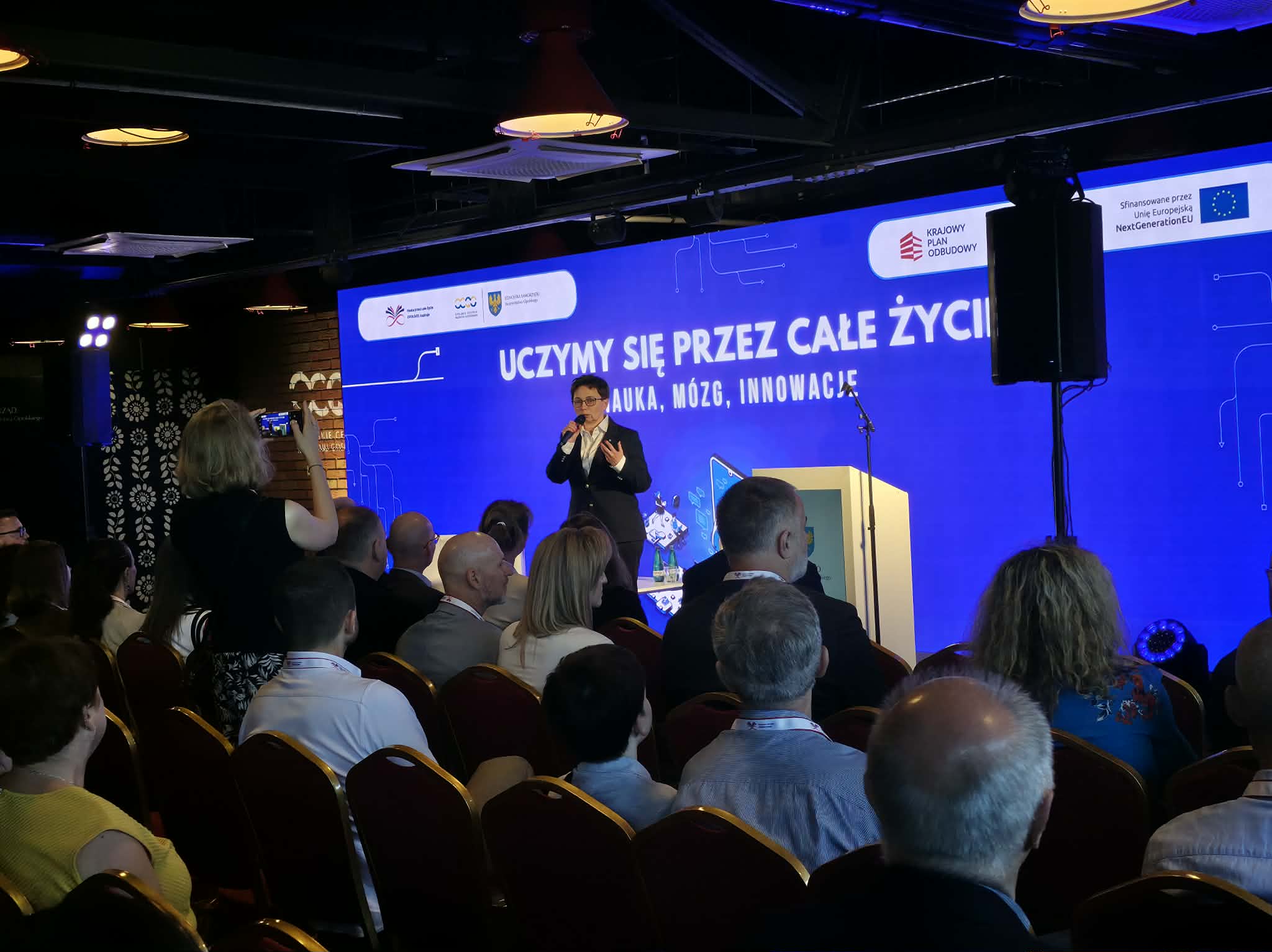 Kobieta stoi na scenie za mównicą i przemawia do mikrofonu podczas oficjalnego wydarzenia. Tło stanowi ogromny niebieski ekran z hasłem „UCZYMY SIĘ PRZEZ CAŁE ŻYCIE! NAUKA, MÓZG, INNOWACJE”. Na pierwszym planie widoczni są uczestnicy konferencji siedzący w rzędach na krzesłach z bordową tapicerką. Po lewej stronie jedna z osób na widowni rejestruje wystąpienie za pomocą telefonu komórkowego.