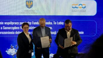 Kobieta i dwóch mężczyzn stoją na scenie, prezentując podpisane dokumenty w ciemnych okładkach. W tle znajduje się niebieski ekran z logotypami Samorządu Województwa Opolskiego oraz Opolskiego Centrum Rozwoju Gospodarki. Widoczny fragment napisu na ekranie dotyczy nawiązania współpracy pomiędzy Fundacją a Samorządem. Zdjęcie zostało zrobione z perspektywy widowni, lekko z dołu.