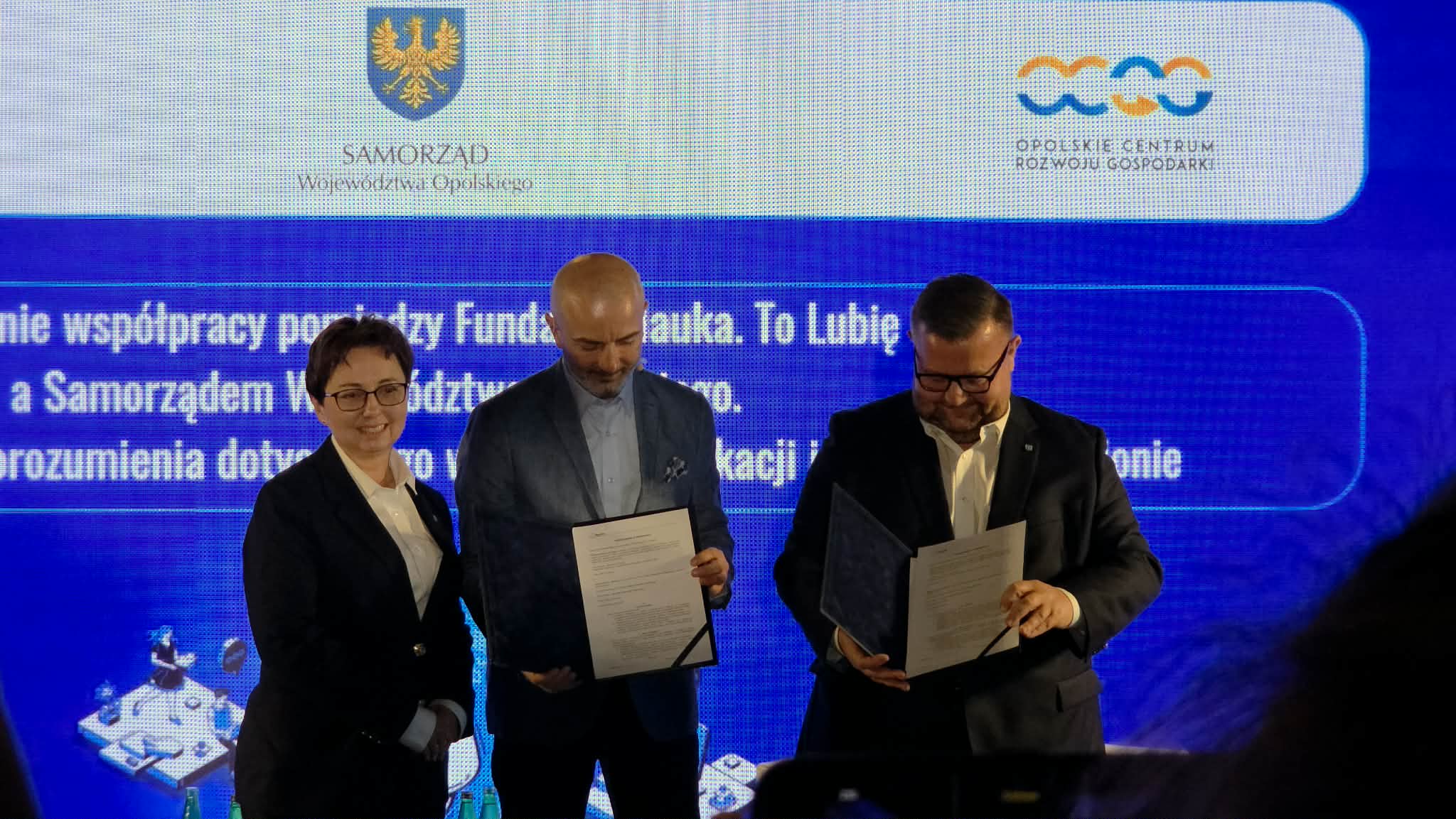 Kobieta i dwóch mężczyzn stoją na scenie, prezentując podpisane dokumenty w ciemnych okładkach. W tle znajduje się niebieski ekran z logotypami Samorządu Województwa Opolskiego oraz Opolskiego Centrum Rozwoju Gospodarki. Widoczny fragment napisu na ekranie dotyczy nawiązania współpracy pomiędzy Fundacją a Samorządem. Zdjęcie zostało zrobione z perspektywy widowni, lekko z dołu.