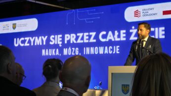 Mężczyzna w ciemnym garniturze i krawacie stoi za białą mównicą z logo Samorządu Województwa Opolskiego i przemawia do mikrofonu. Za nim rozciąga się duży, niebieski ekran z białym napisem „UCZYMY SIĘ PRZEZ CAŁE ŻYCIE. NAUKA, MÓZG, INNOWACJE”. W prawym górnym rogu ekranu widoczne jest logo Krajowego Planu Odbudowy. Na pierwszym planie widać tyły głów kilku osób siedzących na widowni.
