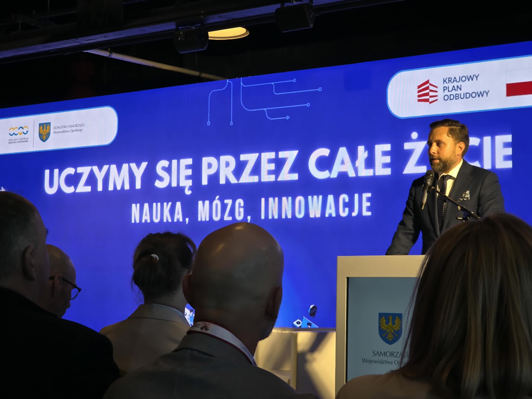 Mężczyzna w ciemnym garniturze i krawacie stoi za białą mównicą z logo Samorządu Województwa Opolskiego i przemawia do mikrofonu. Za nim rozciąga się duży, niebieski ekran z białym napisem „UCZYMY SIĘ PRZEZ CAŁE ŻYCIE. NAUKA, MÓZG, INNOWACJE”. W prawym górnym rogu ekranu widoczne jest logo Krajowego Planu Odbudowy. Na pierwszym planie widać tyły głów kilku osób siedzących na widowni.