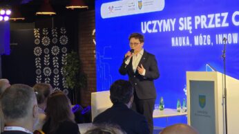 Kobieta w ciemnym garniturze i okularach stoi na scenie, przemawiając do mikrofonu trzymanego w prawej ręce. Po prawej stronie kadru stoi biała mównica z herbem i napisem „SAMORZĄD Województwa Opolskiego”. W tle wyświetla się niebieski ekran z hasłem konferencji oraz mniejszym stolikiem z butelkami wody. Publiczność siedząca przed sceną jest widoczna od tyłu w dolnej części kadru.