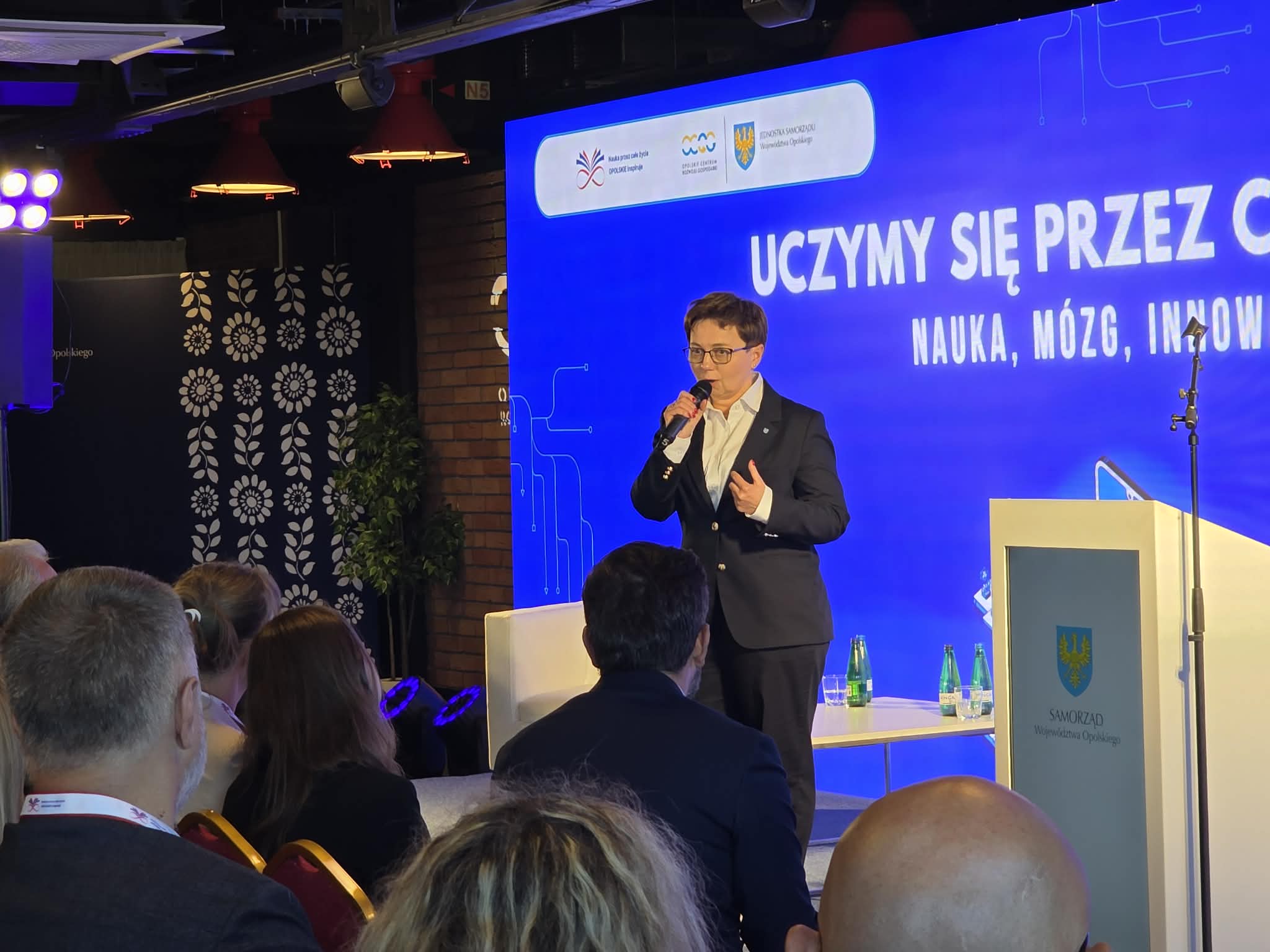 Kobieta w ciemnym garniturze i okularach stoi na scenie, przemawiając do mikrofonu trzymanego w prawej ręce. Po prawej stronie kadru stoi biała mównica z herbem i napisem „SAMORZĄD Województwa Opolskiego”. W tle wyświetla się niebieski ekran z hasłem konferencji oraz mniejszym stolikiem z butelkami wody. Publiczność siedząca przed sceną jest widoczna od tyłu w dolnej części kadru.