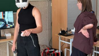 Osoba korzystająca z gogli VR podczas zajęć w sali dydaktycznej, obok nauczyciel obserwuje przebieg ćwiczenia.