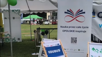 Stoisko edukacyjne na trawie podczas wydarzenia plenerowego. Na pierwszym planie znajduje się niebieski leżak z napisem „EDUBUS”. Obok stoi duża biała tablica promująca projekt „Nauka przez całe życie OPOLSKIE inspiruje” z grafiką otwartej książki i kodem QR. W tle widoczne są namioty wystawowe, balony oraz fragment zabytkowego budynku z wieżami na tle błękitnego nieba.
