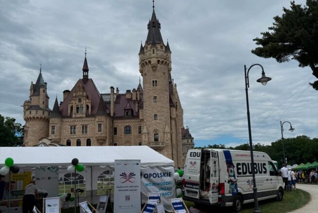 Zdjęcie przedstawia namiot wystawowy ustawiony przed okazałym zamkiem z wysoką wieżą. Obok namiotu zaparkowany jest bus edukacyjny, a wokół znajdują się materiały promocyjne i leżaki dla odwiedzających.