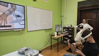 Sala lekcyjna z wyposażeniem do zajęć VR, uczestnik korzysta z gogli wirtualnej rzeczywistości przy stanowisku komputerowym.
