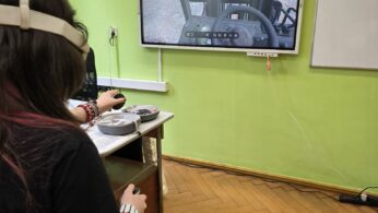 Uczestnik zajęć korzysta z gogli VR, obserwując obraz wyświetlany na monitorze w sali lekcyjnej.