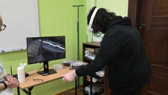 Osoba w goglach VR wykonuje zadanie przy stanowisku komputerowym w sali dydaktycznej.