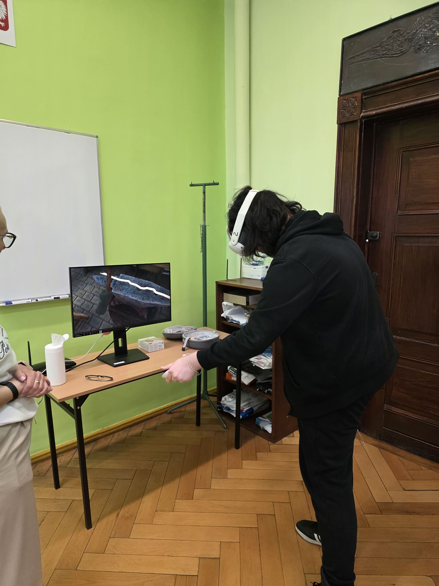 Osoba w goglach VR wykonuje zadanie przy stanowisku komputerowym w sali dydaktycznej.