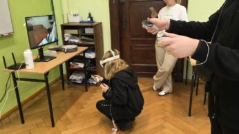 Uczestnik zajęć z goglami VR wykonuje ćwiczenie, a obok znajduje się stanowisko komputerowe i opiekun zajęć.