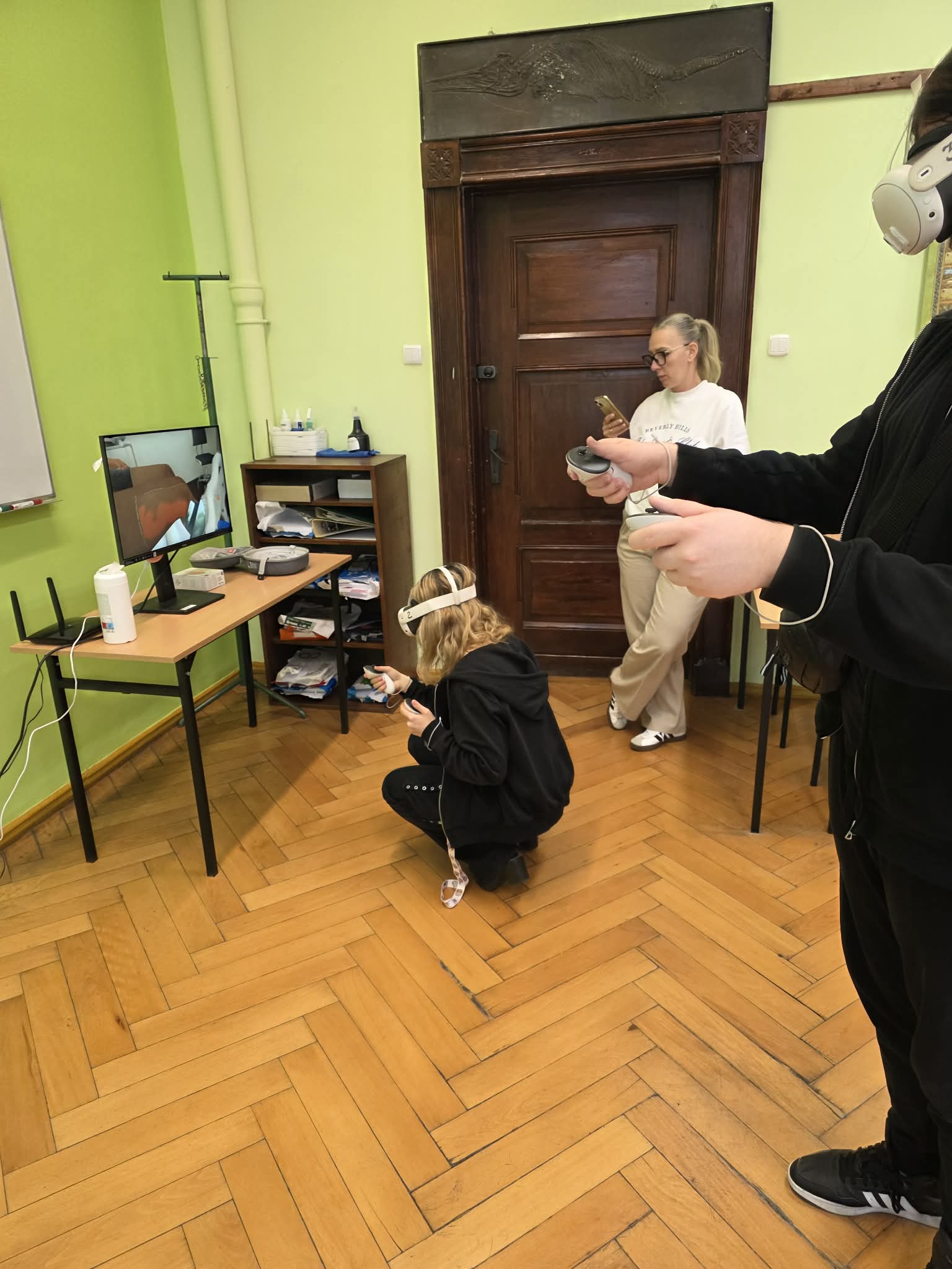 Uczestnik zajęć z goglami VR wykonuje ćwiczenie, a obok znajduje się stanowisko komputerowe i opiekun zajęć.