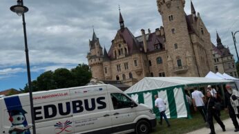 Widok zamku i strefy wydarzenia plenerowego. Na pierwszym planie stoi bus edukacyjny oraz biało-zielony namiot, przy którym poruszają się uczestnicy wydarzenia.