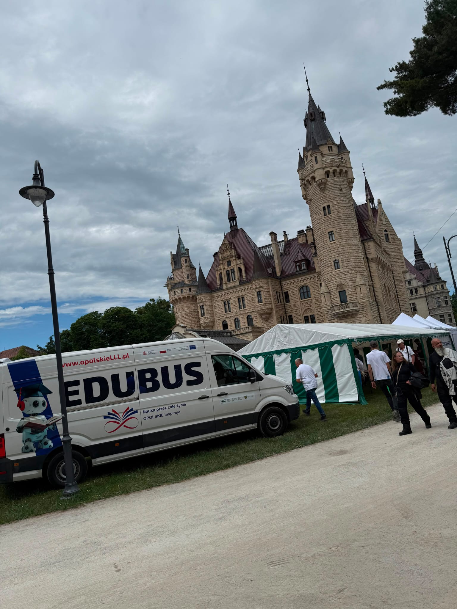 Widok zamku i strefy wydarzenia plenerowego. Na pierwszym planie stoi bus edukacyjny oraz biało-zielony namiot, przy którym poruszają się uczestnicy wydarzenia.