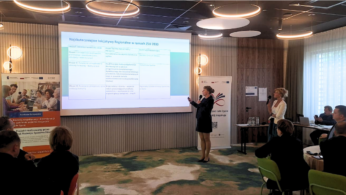 Osoba prowadząca prezentację przed ekranem z wyświetlonym slajdem w sali konferencyjnej.
