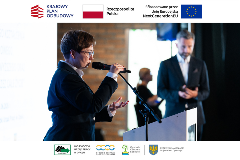 Kobieta w okularach i ciemnej marynarce przemawia do mikrofonu, stojąc za białą mównicą. Z boku widoczny jest fragment jasnego ekranu z tekstem. W tle po prawej stronie stoi rozmyty mężczyzna w garniturze. Zdjęcie ma białe ramki u góry i u dołu z logotypami m.in. Krajowego Planu Odbudowy oraz Wojewódzkiego Urzędu Pracy w Opolu.