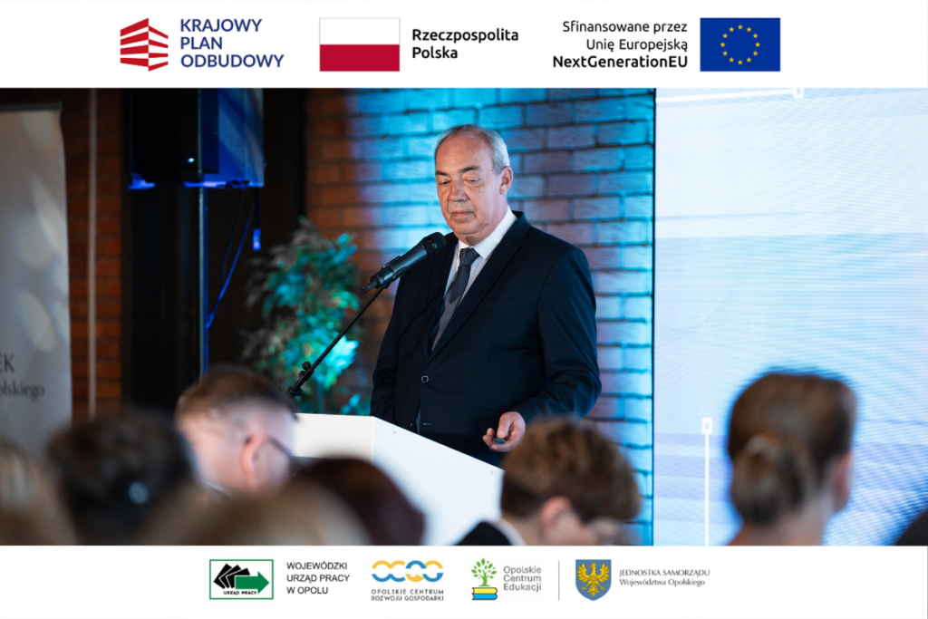 Mężczyzna w ciemnym garniturze i krawacie stoi za białą mównicą, trzymając w dłoni pilota do prezentacji i patrząc w dół. Za nim widać ceglaną ścianę z niebieskim podświetleniem oraz fragment jasnego ekranu multimedialnego. Na pierwszym planie znajdują się nieostre głowy siedzących uczestników wydarzenia. Zdjęcie ma białe ramki u góry i u dołu z kompletem oficjalnych logotypów projektowych.