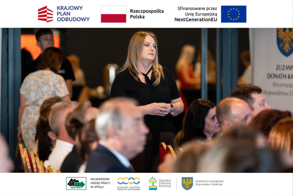 Kobieta w czarnej bluzce z wiązaniem pod szyją stoi na środku sali konferencyjnej ze splecionymi dłońmi, patrząc w stronę sceny. Wokół niej, w dolnej części kadru oraz po prawej stronie, widać rozmyte profile siedzących uczestników konferencji. W tle po prawej stronie majaczy fragment tablicy informacyjnej z logo województwa opolskiego. Kadr otaczają białe pasy z unijnymi i lokalnymi logotypami.