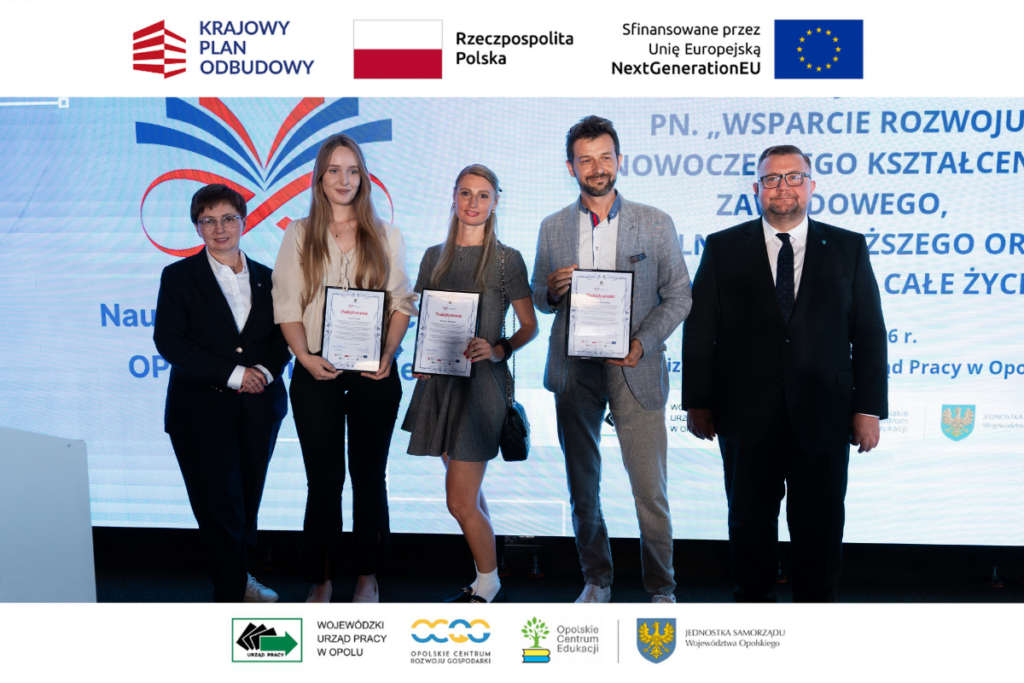 Grupa pięciu osób stoi w rzędzie na scenie przed dużym ekranem z grafiką projektową. Trzy osoby w środku prezentują trzymane w rękach oprawione dyplomy lub podziękowania. Po bokach grupy stoją kobieta w ciemnym garniturze oraz mężczyzna w okularach i czarnym garniturze z krawatem. Wokół kadru umieszczono białe paski z logotypami funduszy europejskich i partnerów regionalnych.