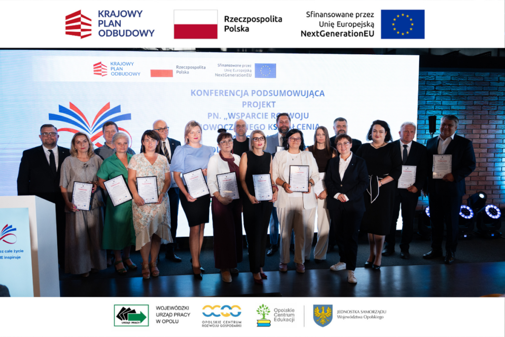 Liczna grupa uczestników konferencji stoi na scenie, pozując do wspólnego zdjęcia na tle dużego ekranu z napisem o podsumowaniu projektu. Wiele osób z przodu trzyma w rękach oficjalne podziękowania w ramkach. W dolnej i górnej części zdjęcia znajdują się białe paski z logotypami identyfikacyjnymi projektu, w tym Unii Europejskiej NextGenerationEU.