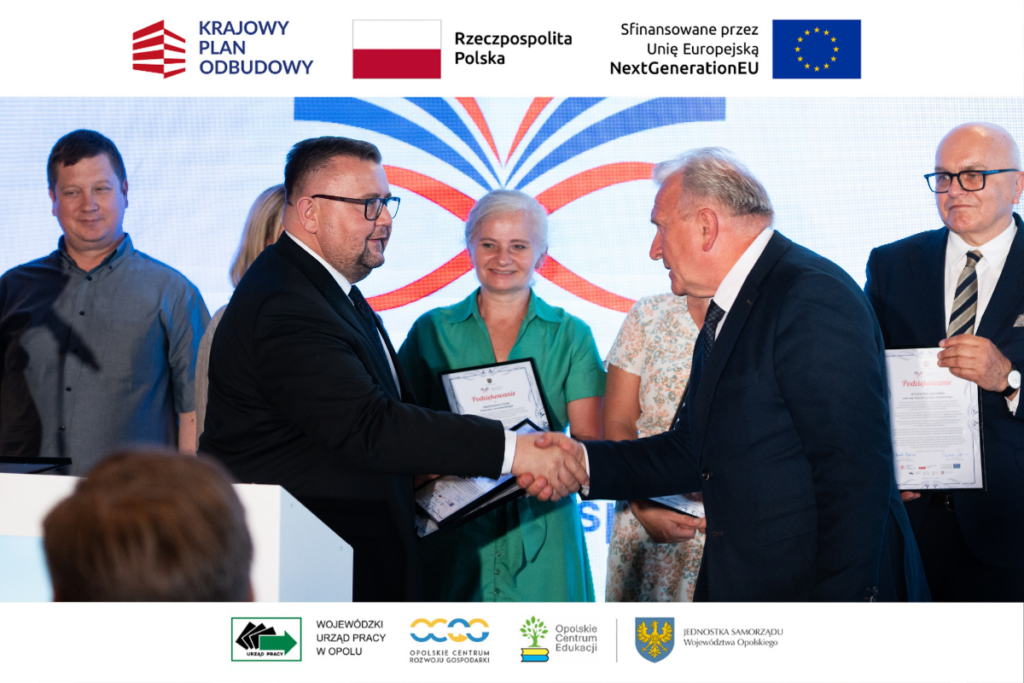Dwaj mężczyźni w garniturach podają sobie ręce w uścisku na tle ekranu z kolorowym logotypem. Mężczyzna po lewej stronie nosi okulary, a po prawej stronie stoi starszy mężczyzna z siwymi włosami. W tle widać uśmiechnięte twarze innych osób trzymających oprawione dokumenty. Obraz posiada białe obramowanie z logotypami instytucji państwowych i samorządowych.