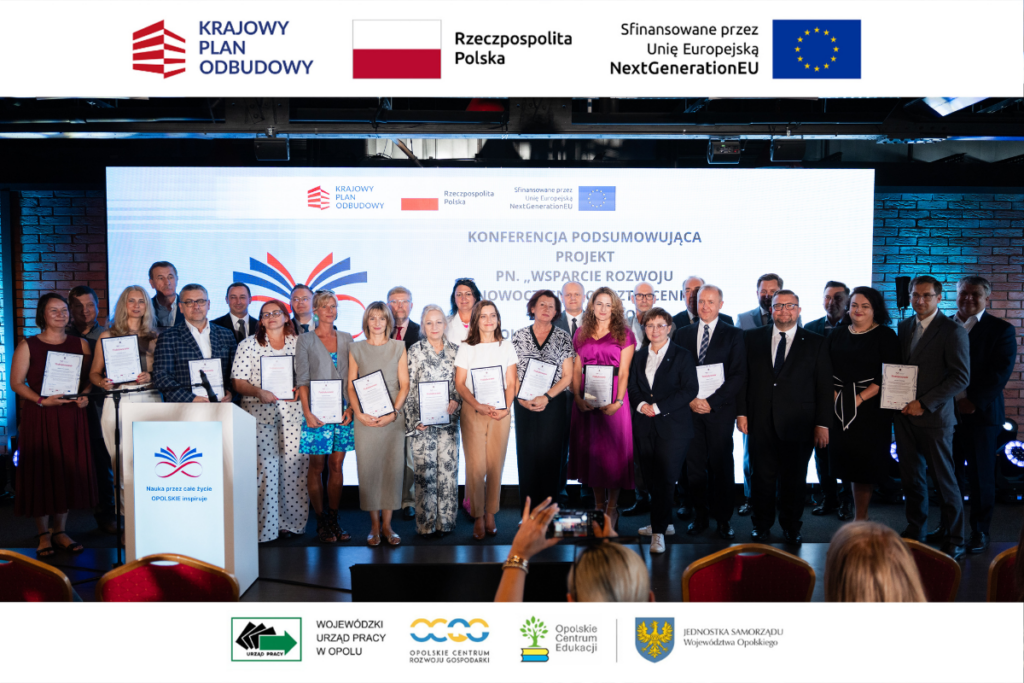 Szerokie ujęcie grupy kilkunastu osób stojących w rzędzie na scenie z dyplomami w rękach przed ekranem z napisem „KONFERENCJA PODSUMOWUJĄCA PROJEKT”. Po lewej stronie widoczna jest biała mównica, a na pierwszym planie w dole kadru widać głowy publiczności oraz uniesioną dłoń ze smartfonem. Zdjęcie otaczają białe pasy z logotypami jednostek samorządu i funduszy pomocowych.