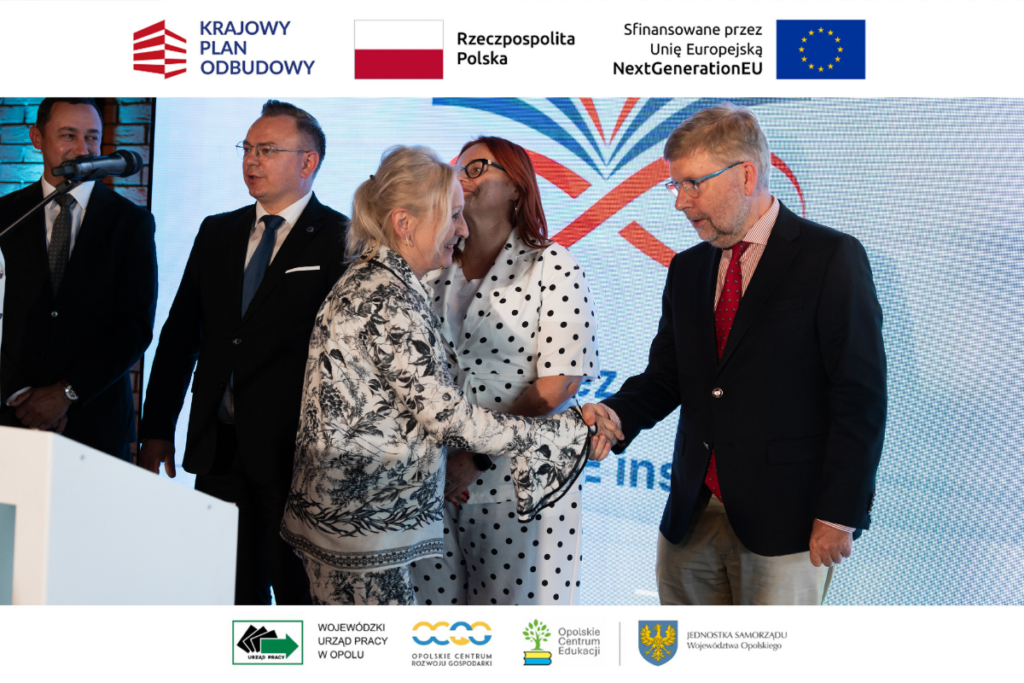 Kobieta w jasnym garniturze w roślinny wzór wita się uściskiem dłoni z mężczyzną w okularach, brodzie i ciemnej marynarce z czerwonym krawatem. W tle stoi inna kobieta w białym garniturze w czarne grochy oraz dwaj mężczyźni w ciemnych garniturach. Kadr zamykają od góry i dołu białe marginesy z logotypami Krajowego Planu Odbudowy i partnerów lokalnych.
