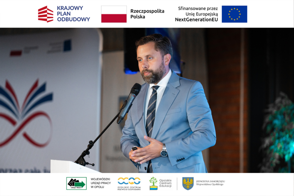 Mężczyzna z zarostem, ubrany w błękitny garnitur, białą koszulę i ciemny krawat w paski, przemawia stojąc za białą mównicą z mikrofonem. W lewej dłoni trzyma mały pilot do prezentacji, a prawą wykonuje gest na wysokości brzucha. W tle po lewej stronie widać fragment planszy z logotypem otwartej książki i znaku nieskończoności. Kadr zamykają białe paski z logotypami m.in. Krajowego Planu Odbudowy i Wojewódzkiego Urzędu Pracy w Opolu.