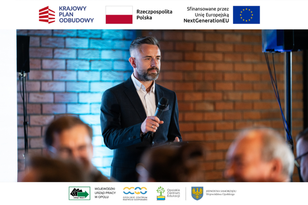 Mężczyzna z krótką brodą i starannie ułożonymi włosami stoi na tle ceglanej ściany, trzymając w dłoni mikrofon i patrząc przed siebie. Ma na sobie ciemną marynarkę i białą koszulę z rozpiętym guzikiem pod szyją. Na pierwszym planie w dolnej części kadru widać rozmyte głowy siedzących tyłem uczestników. Wokół zdjęcia znajdują się białe marginesy z kompletem oficjalnych logotypów programowych.