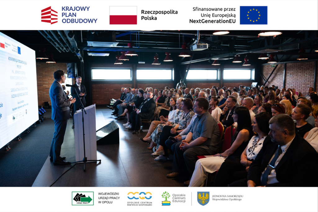 Szerokie ujęcie sali konferencyjnej w industrialnym stylu z ceglanymi ścianami i widocznym pod sufitem czarnym systemem wentylacji. Po lewej stronie, obok białej mównicy i dużego ekranu z tekstem prezentacji, stoi przemawiający mężczyzna w garniturze. Po prawej stronie na krzesłach siedzi kilkadziesiąt osób słuchających wystąpienia. Wokół kadru umieszczono białe paski z logotypami funduszy europejskich oraz partnerów regionalnych.