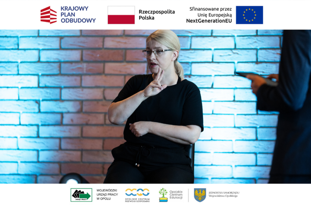 Bliskie ujęcie kobiety w okularach i czarnej bluzce, która siedzi na tle ceglanej ściany oświetlonej niebieskim światłem i tłumaczy wydarzenie na język migowy. Po prawej stronie widać nieostry fragment sylwetki innej osoby trzymającej tablet. Na dole i górze kadru znajdują się białe paski z zestawem oficjalnych logotypów projektowych i partnerskich.