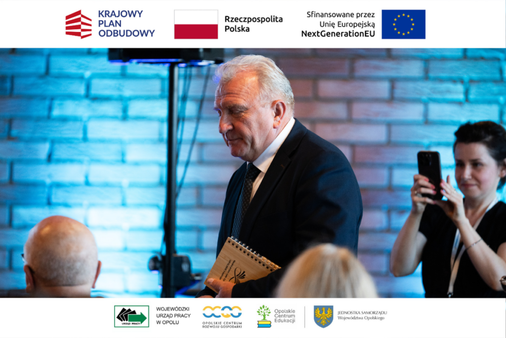 Starszy mężczyzna w ciemnym garniturze i krawacie idzie profilowo przez salę, trzymając w ręku notes z drewnianą okładką. W tle, na tle ceglanej ściany z niebieską iluminacją, widoczna jest kobieta robiąca zdjęcie smartfonem oraz rozmyte głowy innych uczestników konferencji. Fotografia oprawiona jest w białe ramki zawierające logotypy unijne, państwowe oraz regionalne.