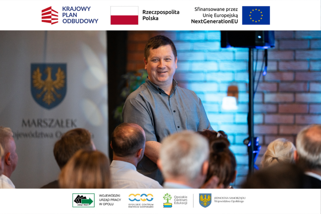 Uśmiechnięty mężczyzna w szarej koszuli stoi przodem do sali, patrząc w stronę widowni, której głowy widoczne są w dolnej części kadru. Po lewej stronie w tle znajduje się baner z godłem województwa opolskiego i napisem „Marszałek Województwa Opolskiego”. Zdjęcie posiada górną i dolną białą belkę z logotypami instytucji i programów finansujących.
