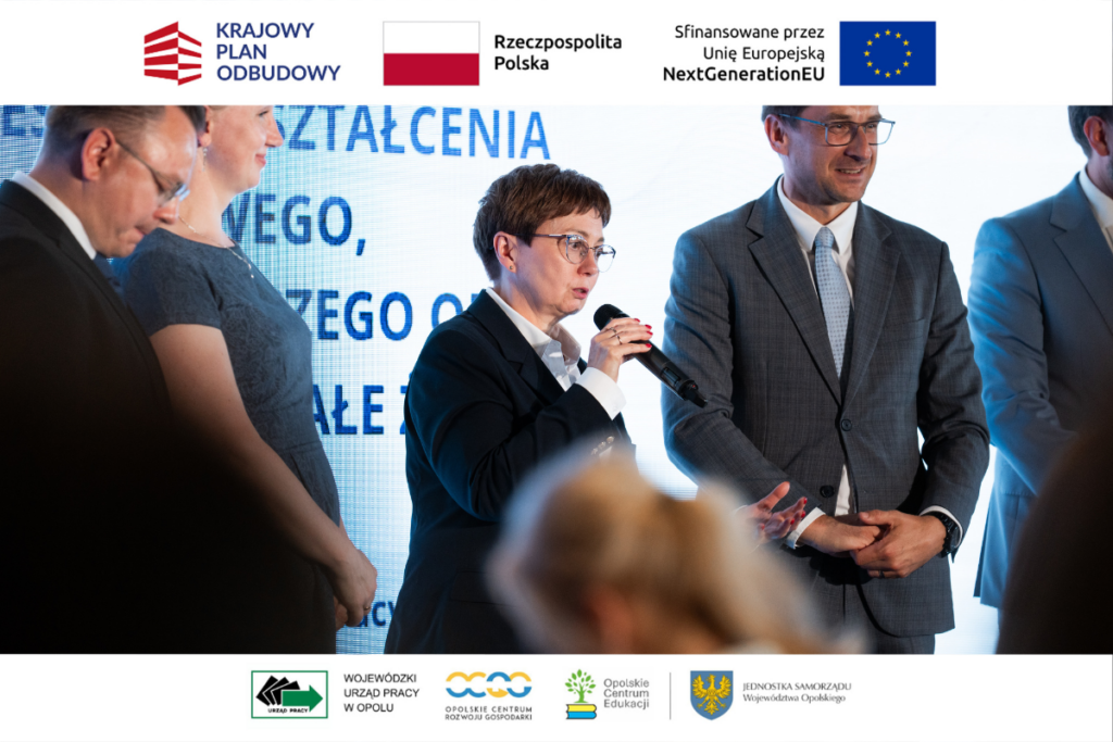 Kobieta w okularach i ciemnym żakiecie przemawia do mikrofonu, stojąc na scenie na tle jasnego ekranu z fragmentami napisów. Obok niej stoją dwaj mężczyźni w garniturach, z których jeden trzyma splecione dłonie. Na pierwszym planie widoczne są nieostre sylwetki publiczności. Górny i dolny margines zdjęcia zawiera oficjalne logotypy organizatorów oraz funduszy europejskich.