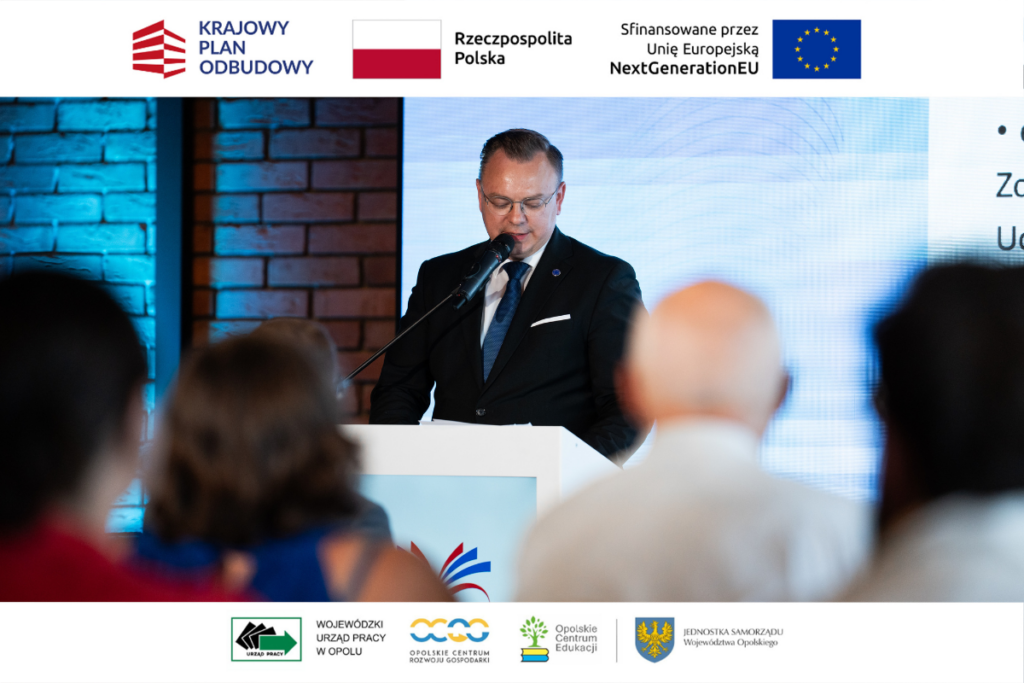 Mężczyzna w okularach i ciemnym garniturze przemawia zza białej mównicy, patrząc w dół na swoje notatki. W tle znajduje się ekran z prezentacją oraz ceglana ściana z niebieskim podświetleniem. Na pierwszym planie widać rozmyte głowy uczestników siedzących tyłem do obiektywu. Wokół zdjęcia umieszczono białe paski z logotypami unijnymi i samorządowymi.