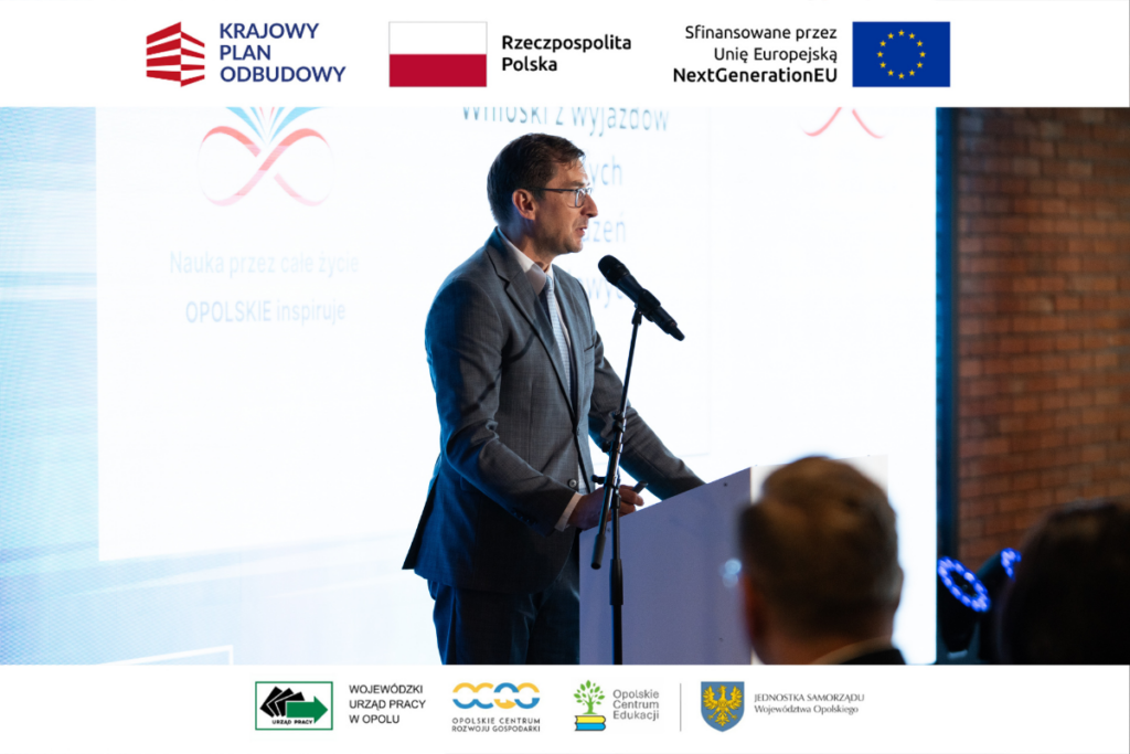 Mężczyzna w okularach i szarym garniturze przemawia do mikrofonu zamontowanego na statywie przy białej mównicy. Prelegent ukazany jest z profilu na tle jasnego ekranu z logotypem i napisem „Nauka przez całe życie OPOLSKIE inspiruje”. Na dole i górze kadru widnieją białe belki z identyfikacją wizualną partnerów i funduszy NextGenerationEU.