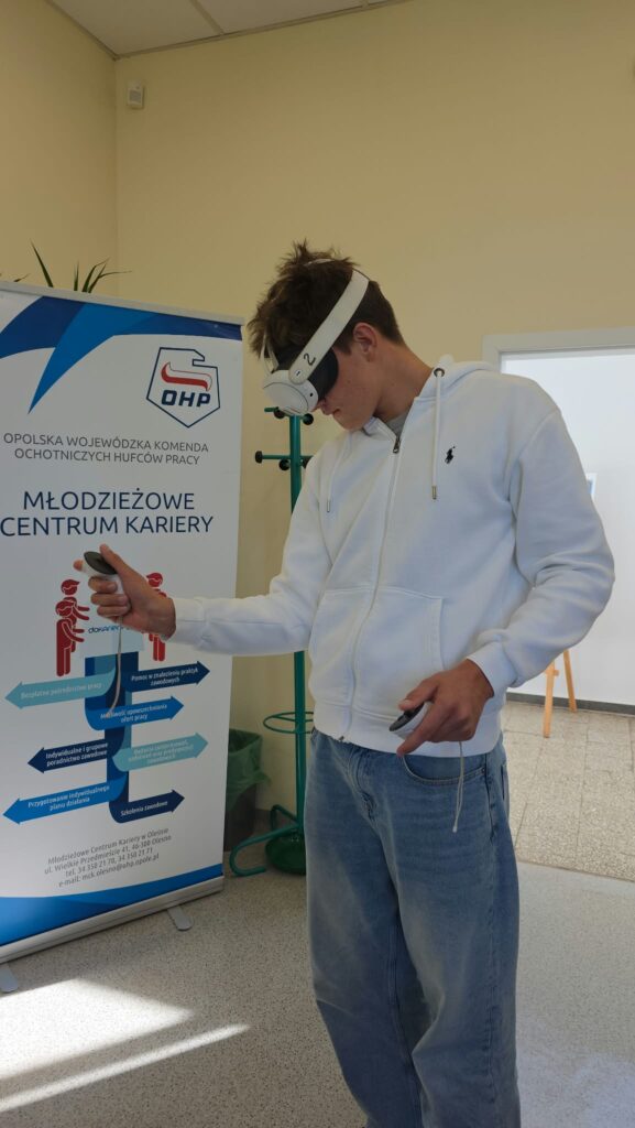 Młody mężczyzna w białej rozpinanej bluzie z kapturem prezentuje gogle VR umieszczone na głowie oraz trzymane w rękach kontrolery. Stoi w widokowym punkcie pomieszczenia tuż obok roll-upu Młodzieżowego Centrum Kariery w Olesnie. Baner przedstawia informacje o bezpłatnym pośrednictwie pracy i doradztwie zawodowym.