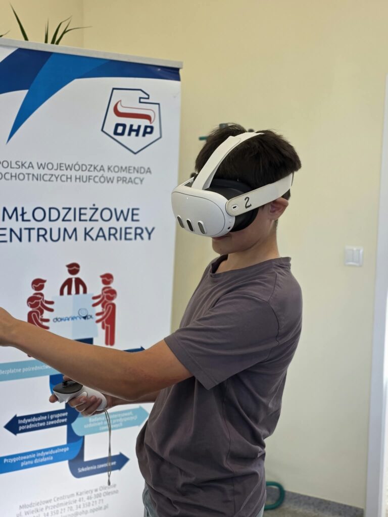Zbliżenie z profilu na chłopca w szarej koszulce, który ma założone białe gogle VR i trzyma w ręce kontroler. W tle znajduje się fragment baneru Młodzieżowego Centrum Kariery OHP z widocznym logo. Chłopiec jest skupiony na interakcji z wirtualnym środowiskiem.