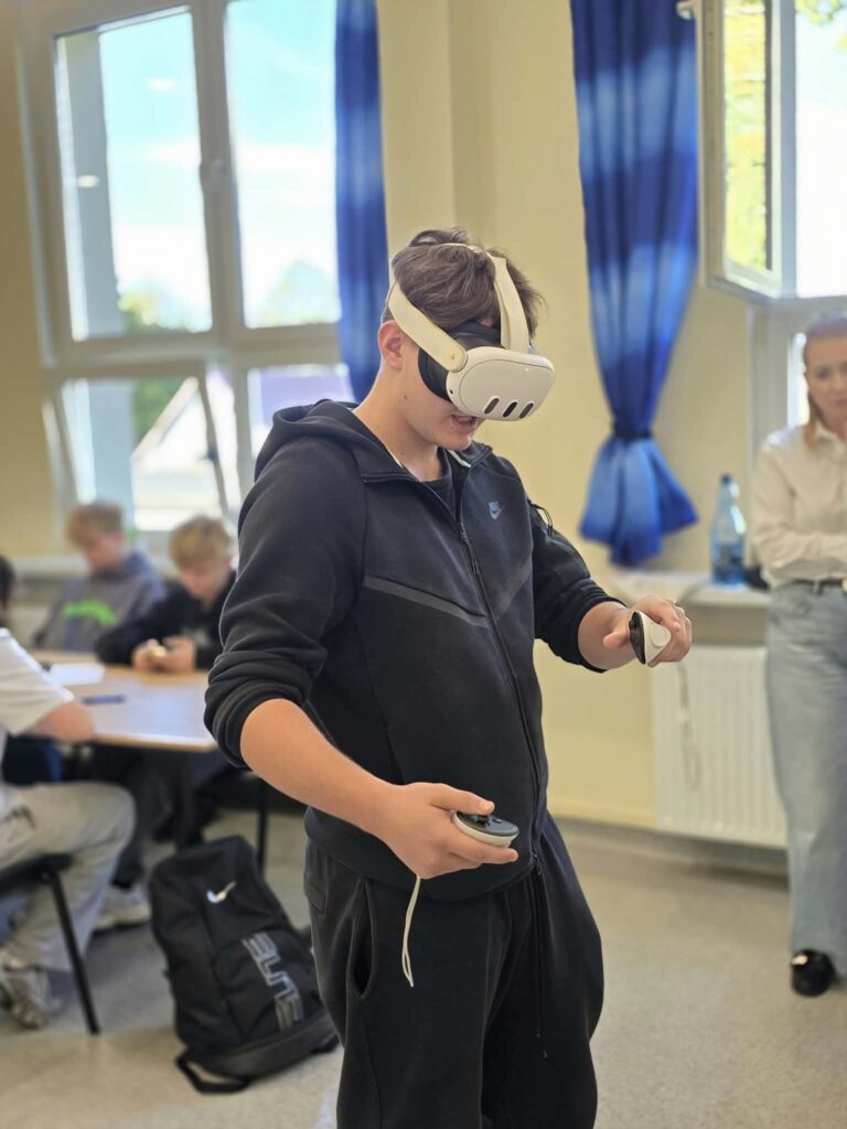 Uczeń w czarnej rozpinanej bluzie i z goglami VR na głowie manipuluje kontrolerami w dłoniach. W tle na drugim planie siedzą inni uczniowie przy stołach, a z boku przygląda im się dorosła osoba. Zajęcia odbywają się w oświetlonym pomieszczeniu z niebieskimi akcentami dekoracyjnymi.