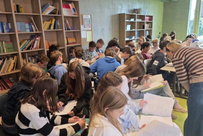 Przepełniona sala biblioteczna, w której młodzież siedzi blisko siebie i wypełnia arkusze papieru. Po prawej stronie prowadząca w paski nachyla się nad uczniami, pomagając im w pracy. W tle znajdują się wysokie regały wypełnione książkami oraz podręcznikami.