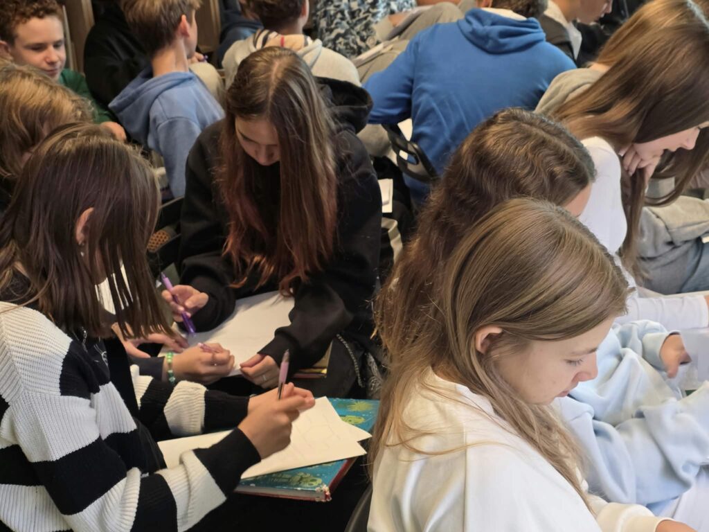 Kadr z bliska przedstawiający grupę dziewcząt i chłopców piszących długopisami na kartkach. Uczniowie skupiają się na wykonywaniu zadań, trzymając podkłady i książki na kolanach. Dziewczyna w czarno-białym swetrze w paski pisze na kartek po lewej stronie kadru.