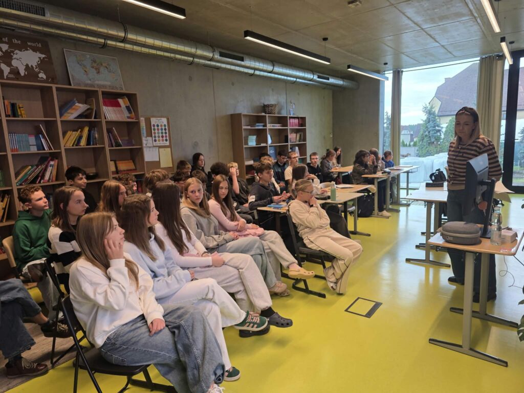 Grupowe zdjęcie uczniów siedzących na krzesłach w nowoczesnej sali lekcyjnej i słuchających nauczycielki prowadzącej zajęcia. Prowadząca stoi przy biurku obok laptopa i ekranu monitora, przekazując wiedzę młodzieży. W tle widać regały pełne książek i materiałów dydaktycznych oraz duże przeszklenie wychodzące na zewnątrz. Pomieszczenie wyróżnia się jasną, zielonkawą podłogą i widocznymi rurami wentylacyjnymi pod sufitem.