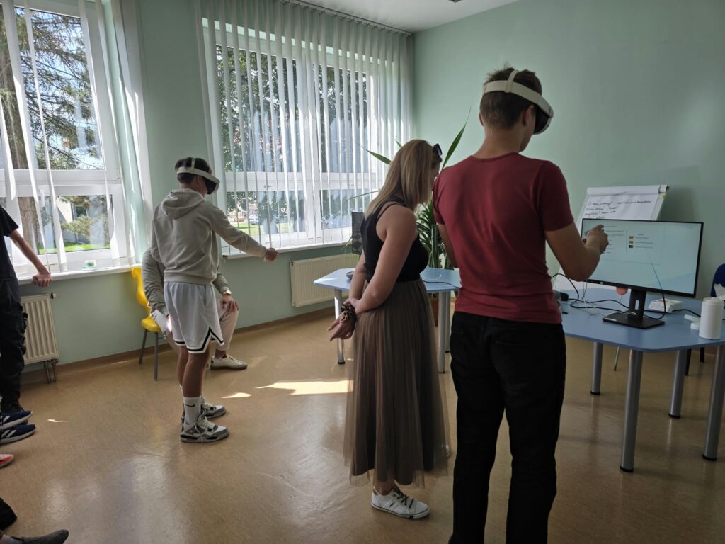 Dwoje młodych ludzi korzysta z gogli rzeczywistości wirtualnej (VR) w jasnej sali lekcyjnej z pionowymi żaluzjami w oknach. Chłopak w czerwonej koszulce stoi tyłem, obserwując interaktywny obraz widoczny na monitorze stacjonarnym. Obok stoją dziewczyna w ciemnej bluzce i spódnicy oraz drugi chłopak wykonujący gesty w goglach VR.