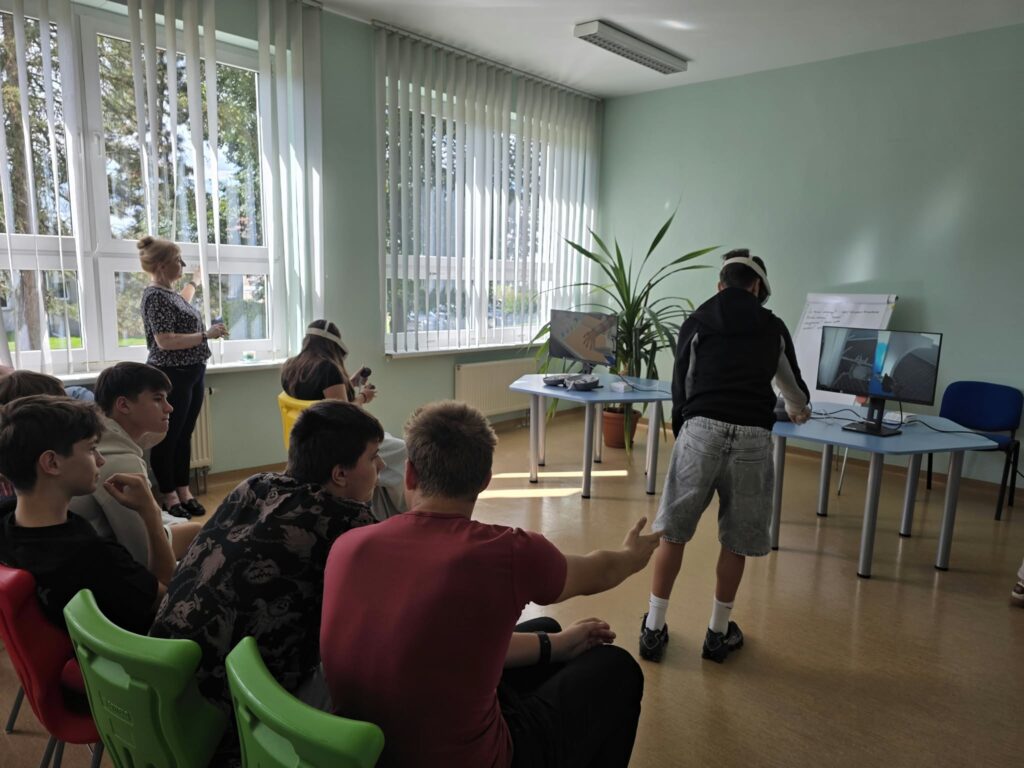 Uczniowie siedzą na kolorowych krzesłach w sali lekcyjnej i obserwują kolegę korzystającego z gogli rzeczywistości wirtualnej. Chłopak w goglach VR stoi przed monitorem, na którym wyświetla się obraz z jego perspektywy. W tle kobieta stoi przy oknie z pionowymi żaluzjami, a w sali znajduje się również roślina doniczkowa